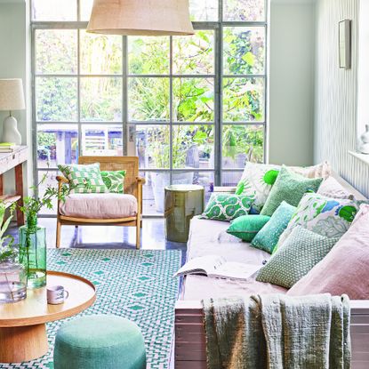 garden room with handmade sofa, green patterned cushions, pink cushions, rattan chair, green patterned indoor/outdoor rug, wooden round coffee table, glass vases, green footstool, lamps, pale green walls