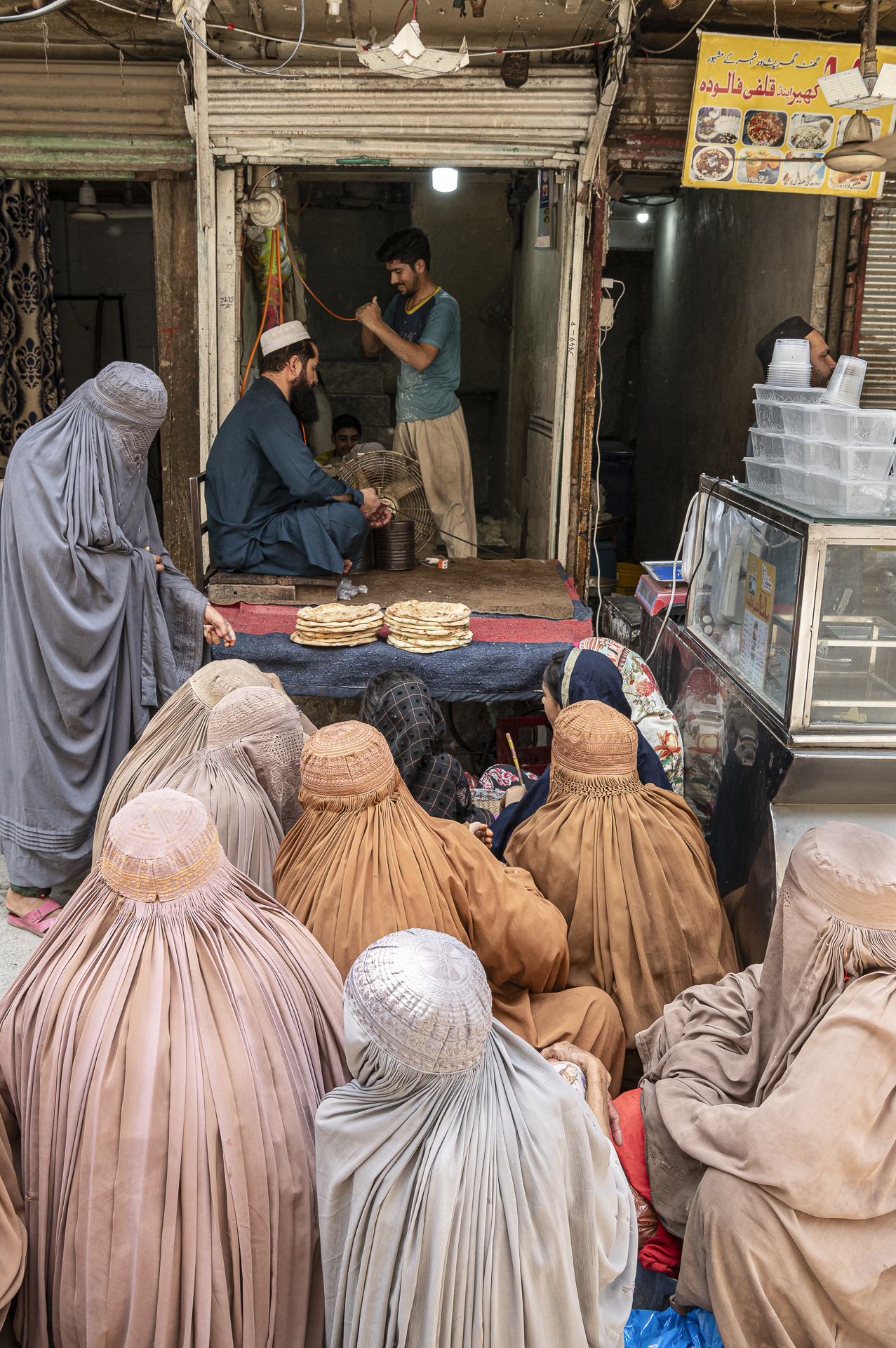 A group of women in traditional attire gathered outside a food stall, watching a vendor prepare bread in a busy market setting