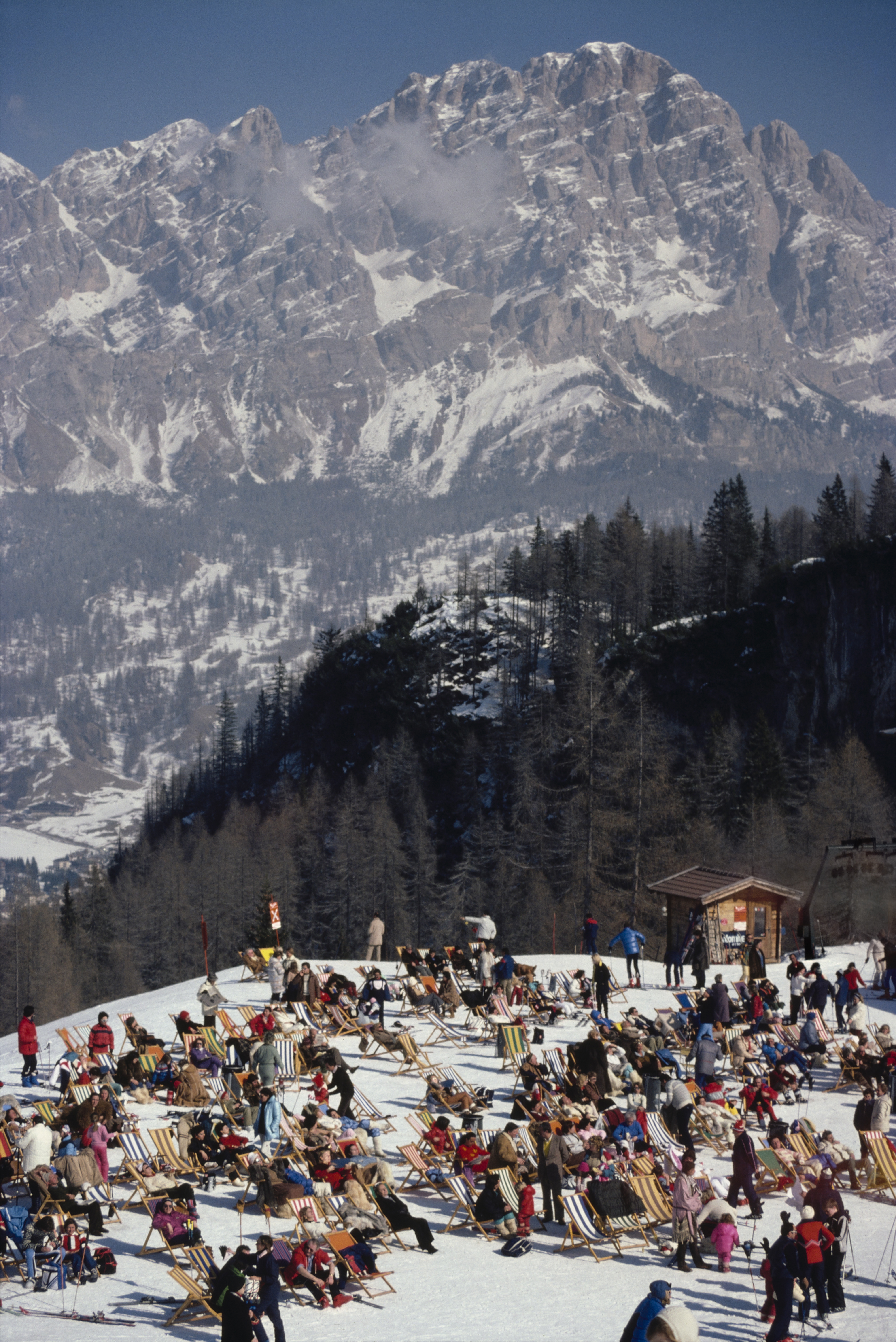 A sunlit alpine plateau filled with rows of deckchairs, where crowds relax in the snow beneath towering mountain cliffs.