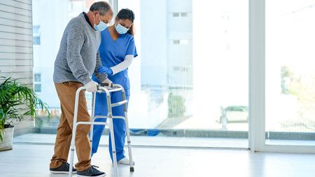 Nurse helping older gentleman use a walker; both wearing surgical masks