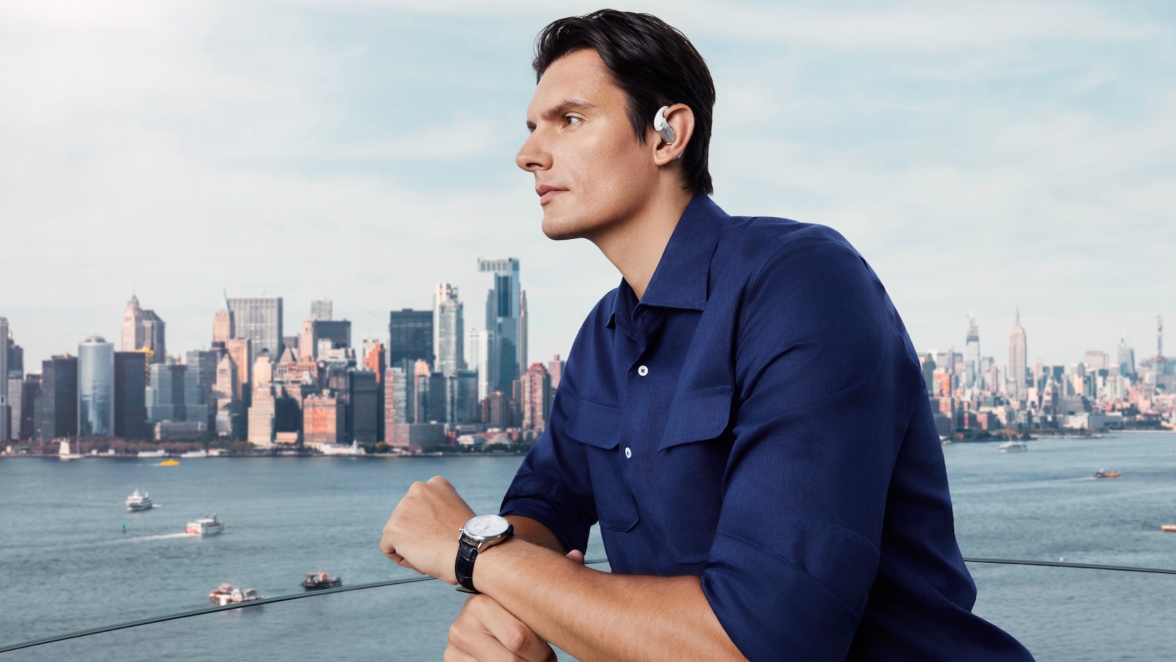 A man in a blue shirt wearing the Shokz OpenFit Pro and standing in front of a city skyline.