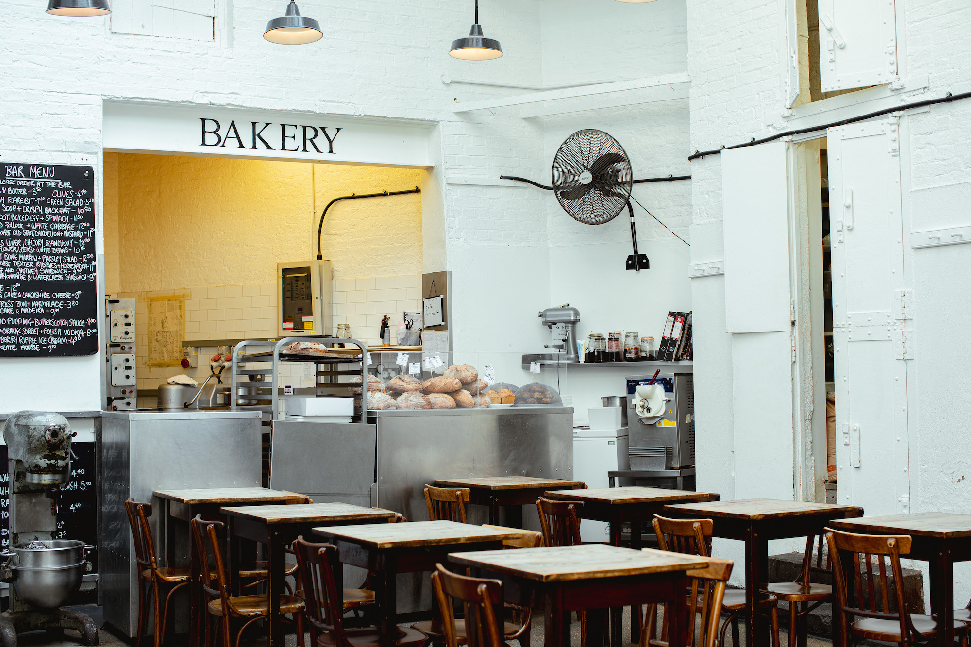 An atmospheric restaurant with essential bistro-style wooden furniture, pendant industrial light, and a sign that reads &quot;BAKERY&quot; in black on white-washed brick walls.