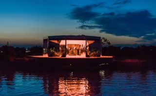 An illuminated gazebo with plants inside situated on a lake.