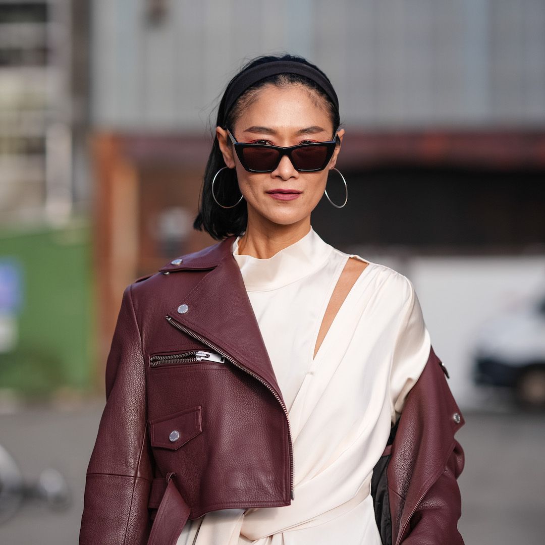 Pornwika Spiecker wears sunglasses, a burgundy leather biker jacket , a white gathered long dress and contoured makeup