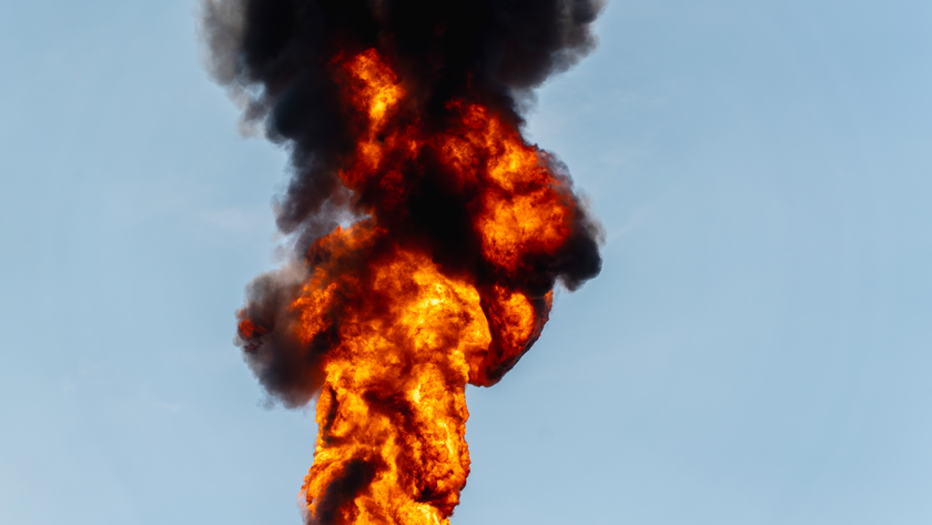 A massive industrial plant surrounded by towering machinery and pipelines, with flames shooting skyward from a flare stack, casting an intense glow against the sky.