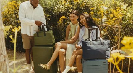 Three friends dressed for a summer trip smile and laugh surrounded by sun-baked greenery and yellow flowers while sitting or standing next to their suitcases.