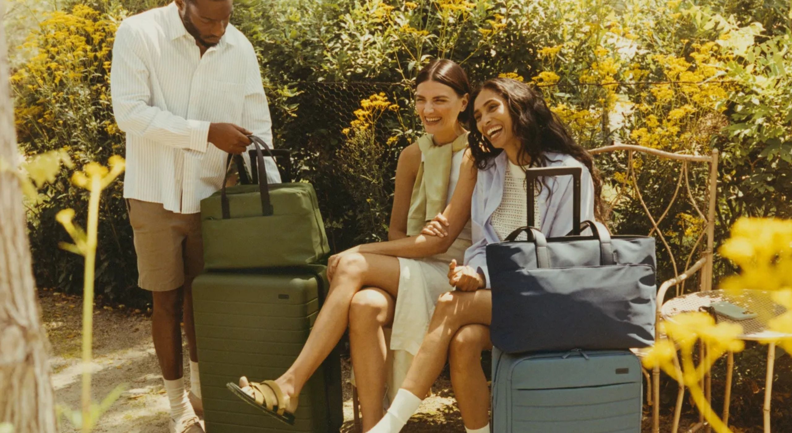 Three friends dressed for a summer trip smile and laugh surrounded by sun-baked greenery and yellow flowers while sitting or standing next to their suitcases.