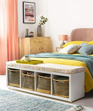 Bedroom with white walls, double bed with yellow and green bedding, stool with storage baskets at the end of the bed.
