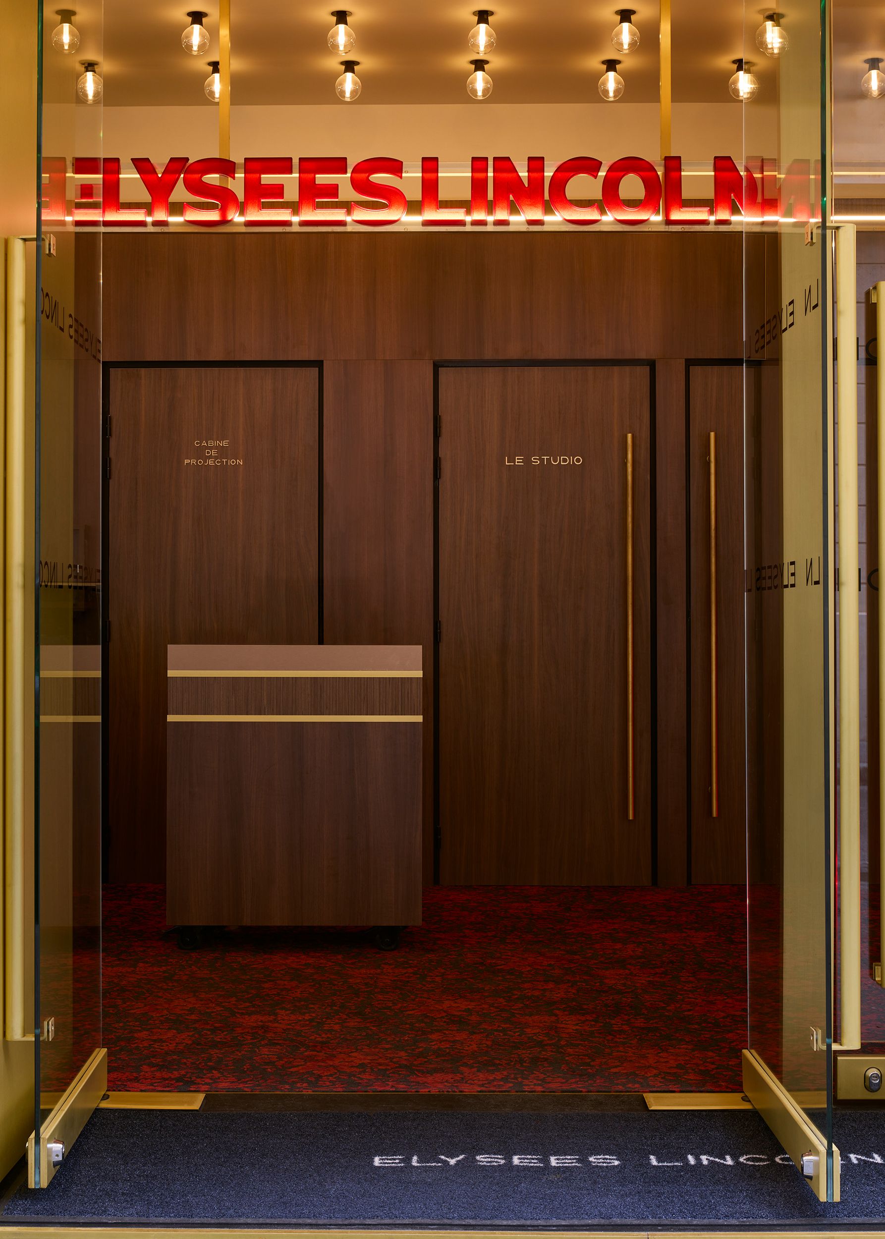 A series of interior shots depicting the retro-futuristic decor of a cinema captures velvet-clad rooms with a palette of reds, pinks, and pale dark greens, theatrical doorways, either arched or draped in curtains, ceiling porthole windows, and a vintage, golden entrance with red neon lights.