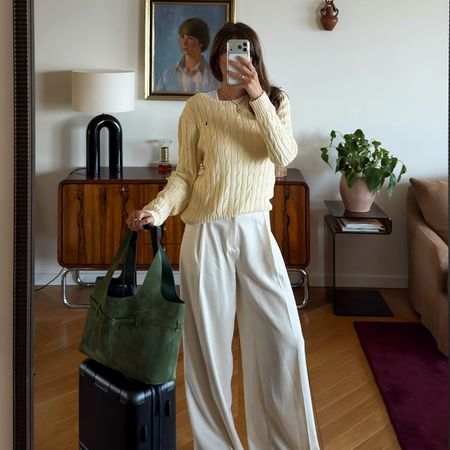 A French woman wearing a butter yellow sweater with cream pants with a suitcase and green suede tote next to her