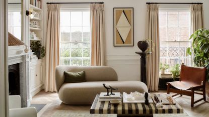 A bright living room with large windows, cream window treatments, a beige couch, a leather chair, a pedestal with a vase, a striped marble table, a fireplace, and white cabinets