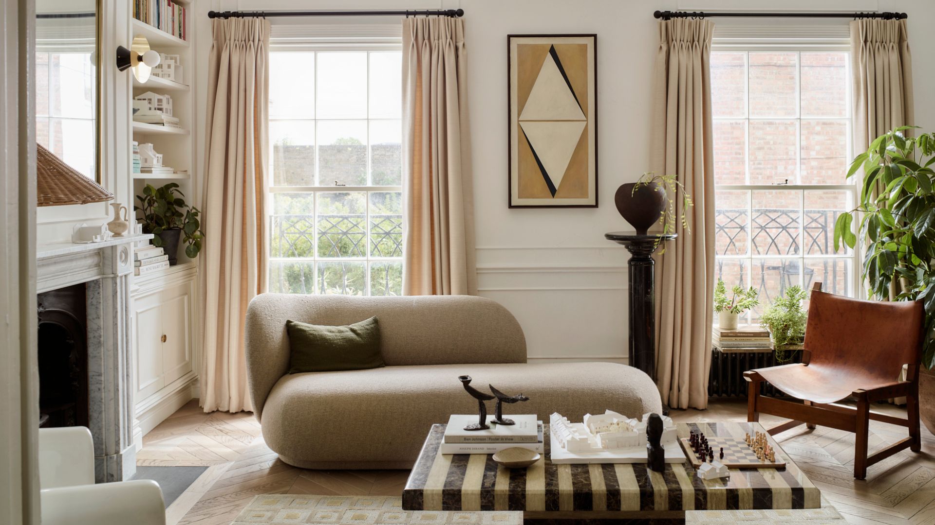 A bright living room with large windows, cream window treatments, a beige couch, a leather chair, a pedestal with a vase, a striped marble table, a fireplace, and white cabinets