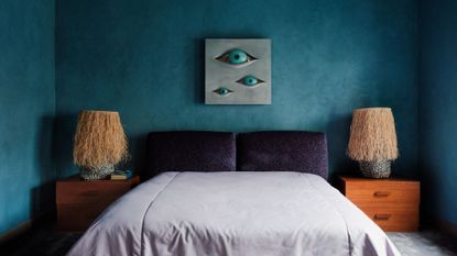 A blue bedroom with eccentric artwork featuring a trio of eyes above a bed with a lilac duvet and a pair of patterned purple pillows beside wood nightstands and table lamps with textured shades