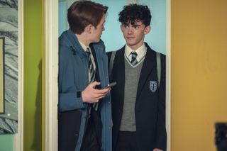 Nick (Kit Connor) and Charlie (Joe Locke) literally bump into each other while walking through a doorway into the classroom, with Nick holding his phone in his hand. Both boys are wearing school uniform and Nick has a blue coat on over the top of his.