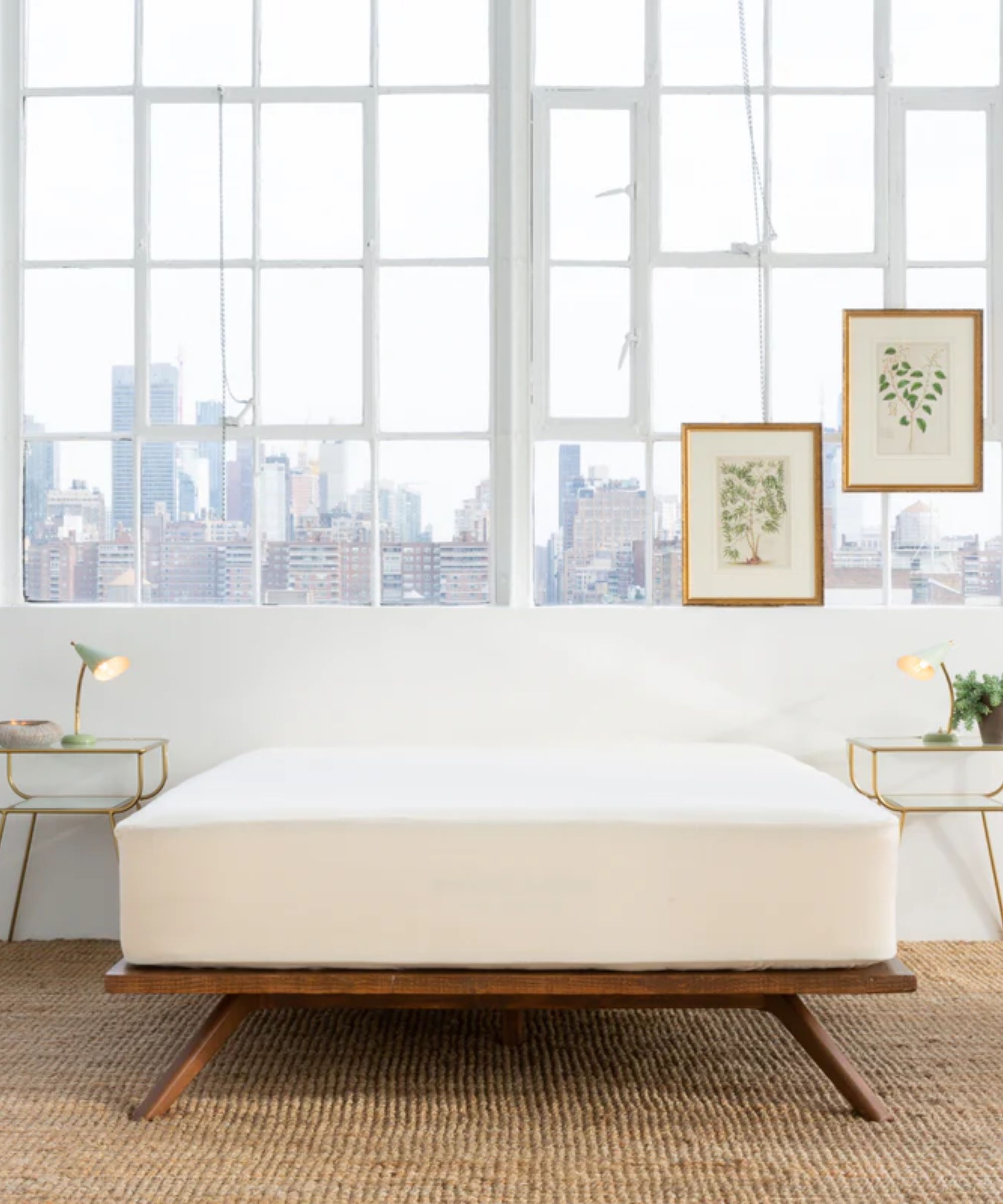 A white Avocado waterproof mattress protector on a low wooden bedframe, without a headboard. The carpet is woven jute, and to either side of the bed are gold bedside tables with matching white lamps, a small grey bowl with a gold interior, and a houseplant. Behind the bed is a white painted wall, and above it are large white crittal windows, with two botanical artworks in gold frames, and a city skyline seen beyond.