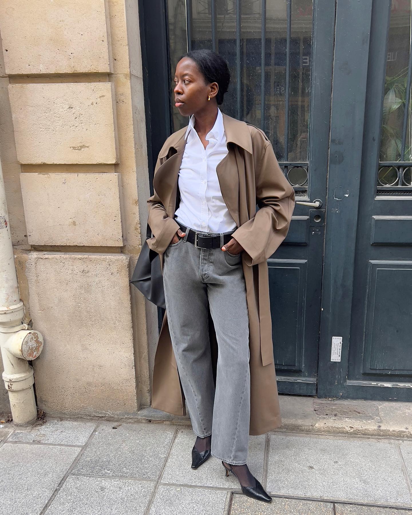 fashion influencer Sylvie Mus standing on the streets of Paris in a trench coat, button-down shirt, gray jeans, and black slingback heels