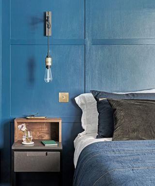 An industrial style exposed bulb hanging wall light attached to a blue paneled wall behind a blue and white bed