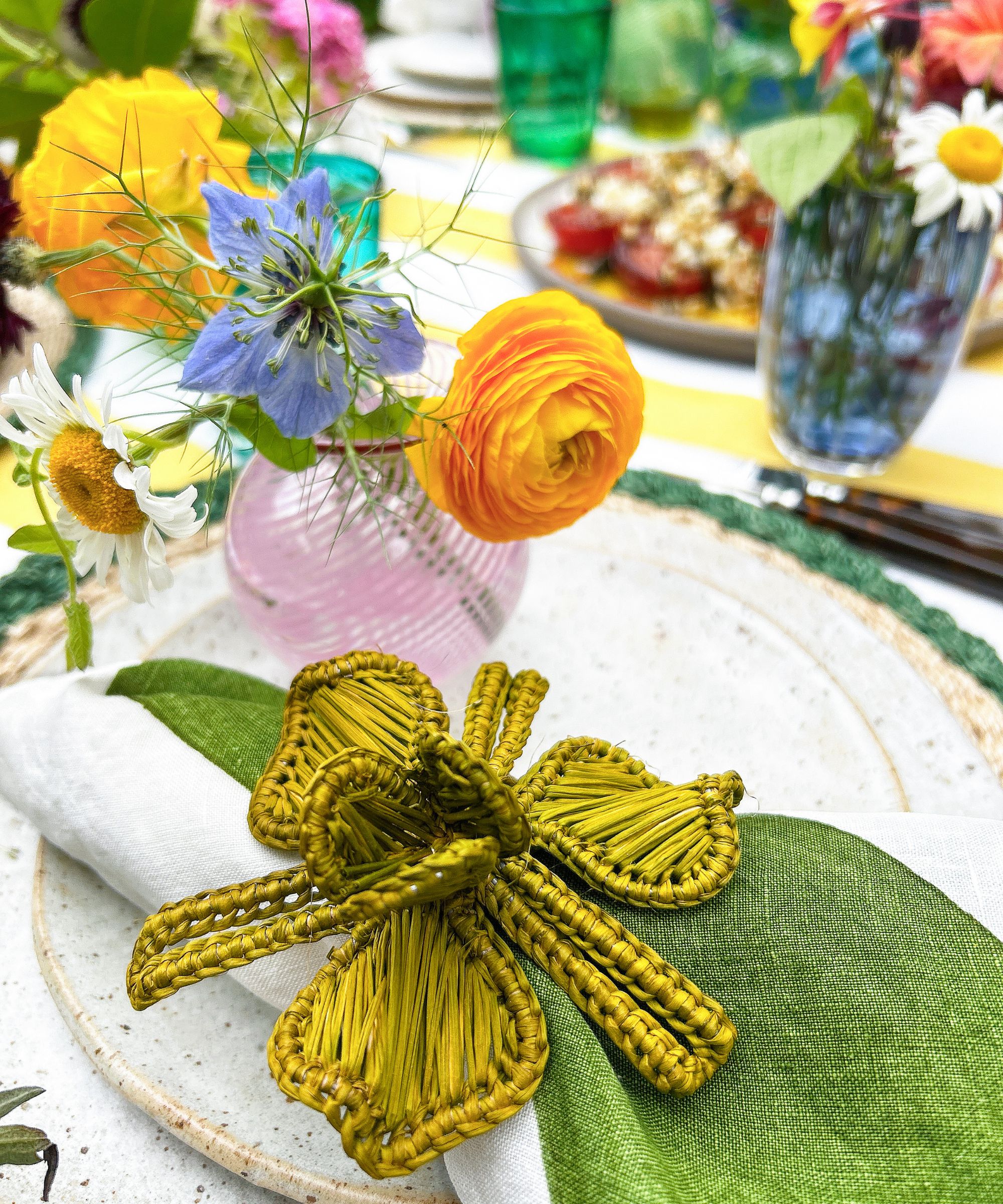 Close up shot of a place setting with a green raffia flower napkin ring on speckled plate with a pink vase of orange and blue flowers and other colorful accessories in the background