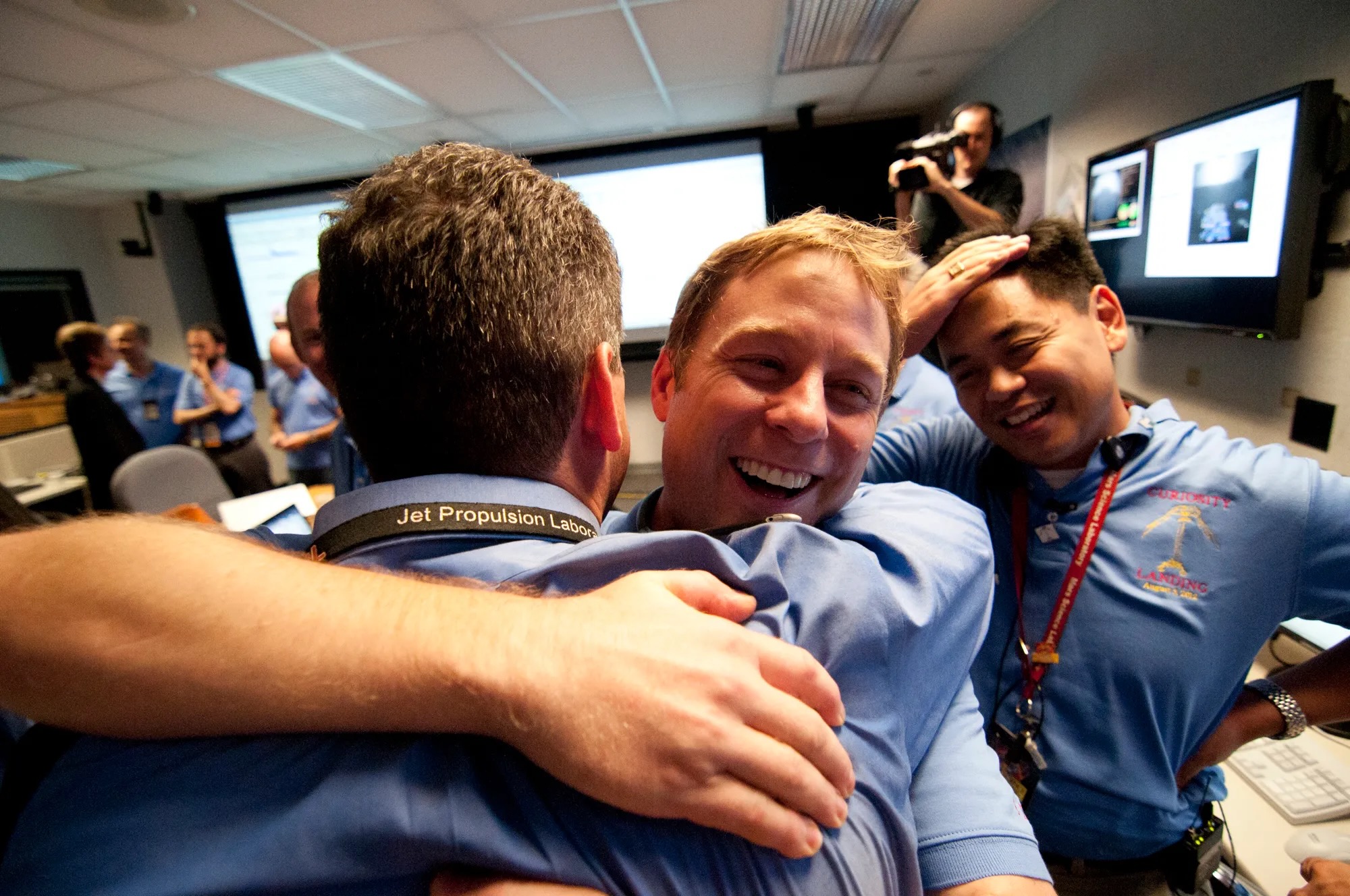 closeup of two smiling men in blue shirts hugging, while other people in the background smile