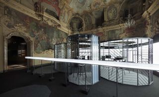 A closer look at the clock-making workshop set up in the Milan’s Palazzo Visconti. The workshop is surrounded by harsh reflector lights, while the rest of the space is dark. The workshop consists of an open framework of interlocking circular spaces.