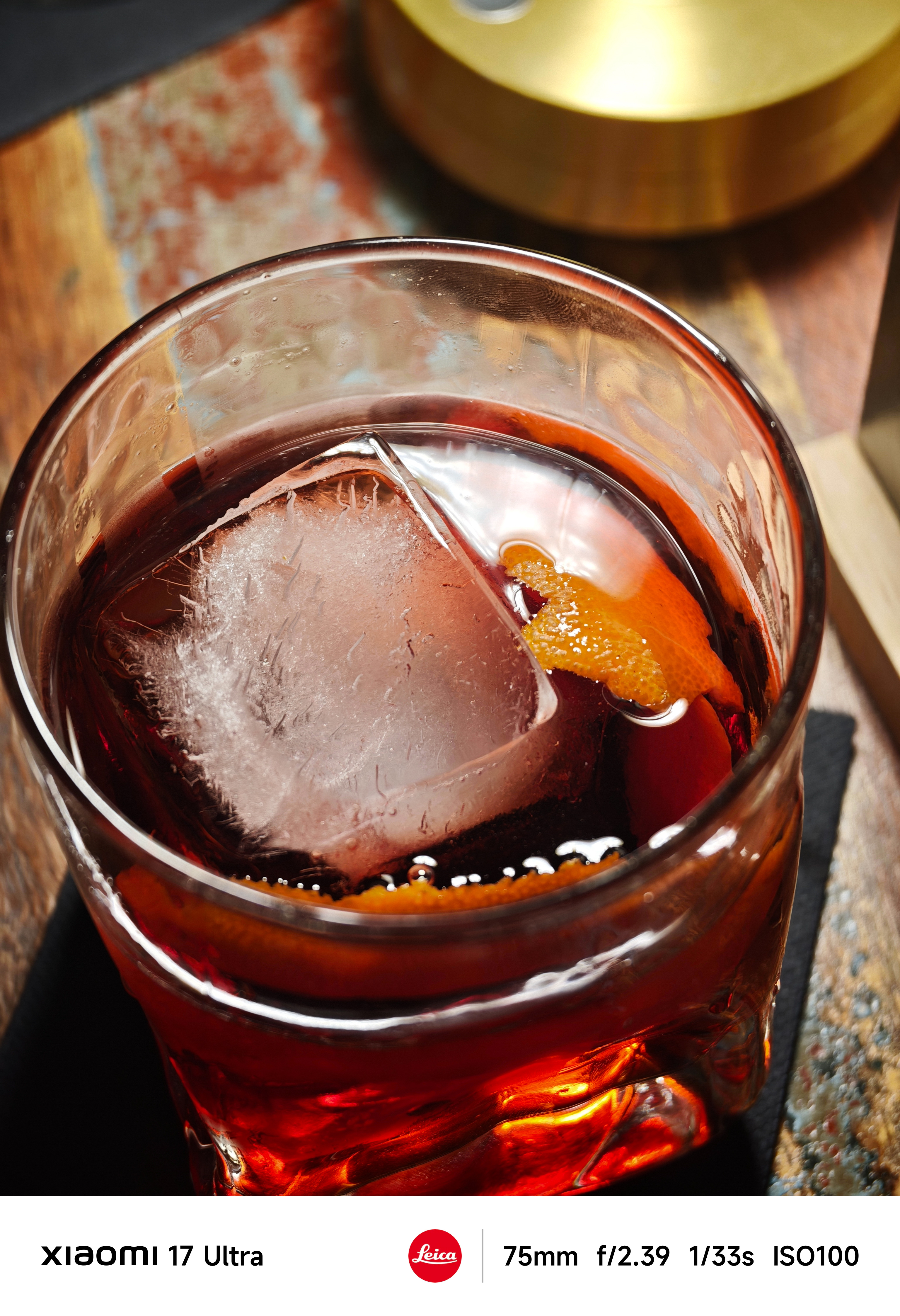 Close-up of a red cocktail in a glass with a large clear ice cube and orange peel garnish, resting on a wooden table.