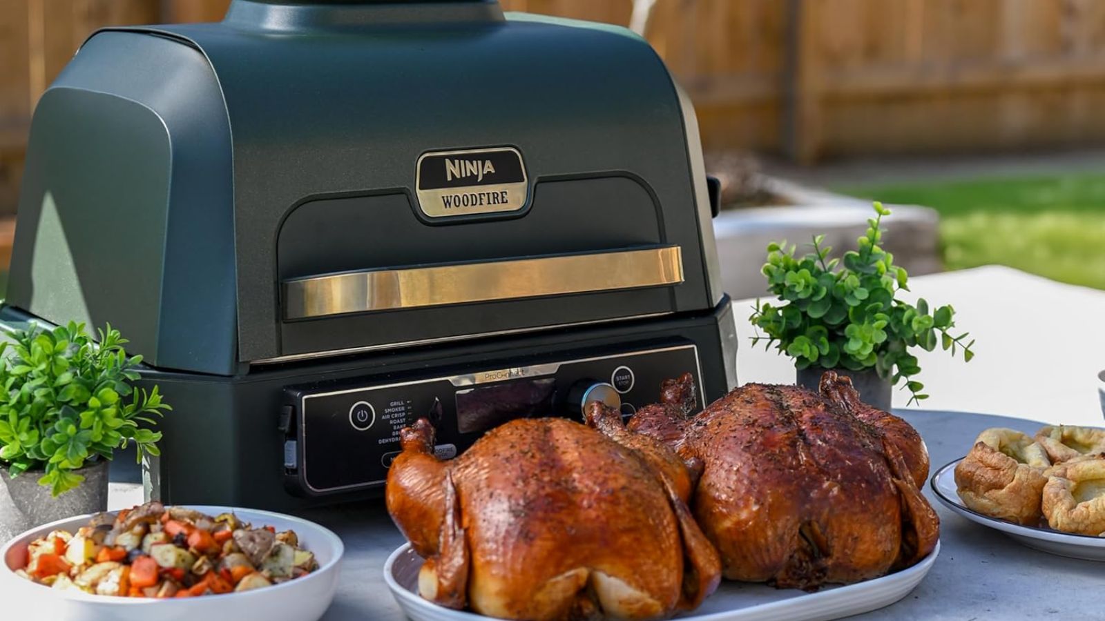 The blue and gold Ninja Woodfire Pro electric grill on an outdoor table surrounded by fried chickens, sides, and potatoes 