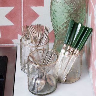 Glass jars used to hold kitchen utensils and cutlery on a white kitchen worktop with pink geo tiled splashback behind
