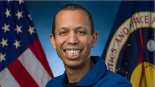 A man smiles with all his teeth wearing a blue collared shirt in front of a blue backdrop with an american flag drooped on the left, and nasa flag dropped on the right.