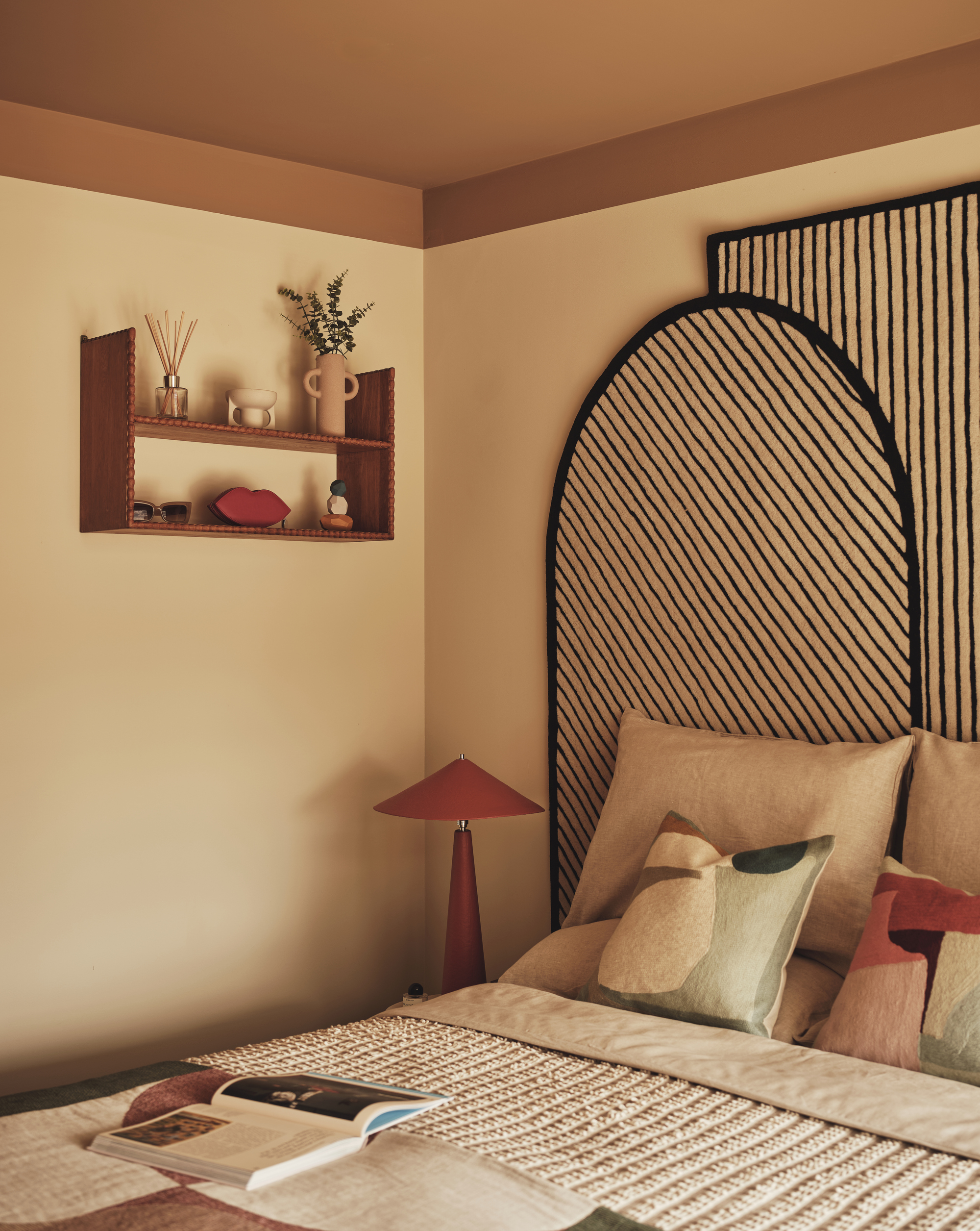 bedroom with subtle yellow walls, orange ceiling, striped headboard, terracotta table lamp, patterned bedding with open book on it, timber shelves with diffuser and vase of flowers