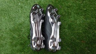 A pair of black football boots lay next to eachother with the silver underside facing upwards on green artificial grass. Sudu SFS FG 01+ Pro