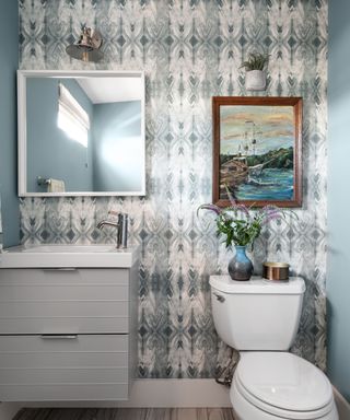 A blue bathroom with a blue and white patterned wallpaper with a mirror and beach wall art, a white sink and toilet, and blue walls either side