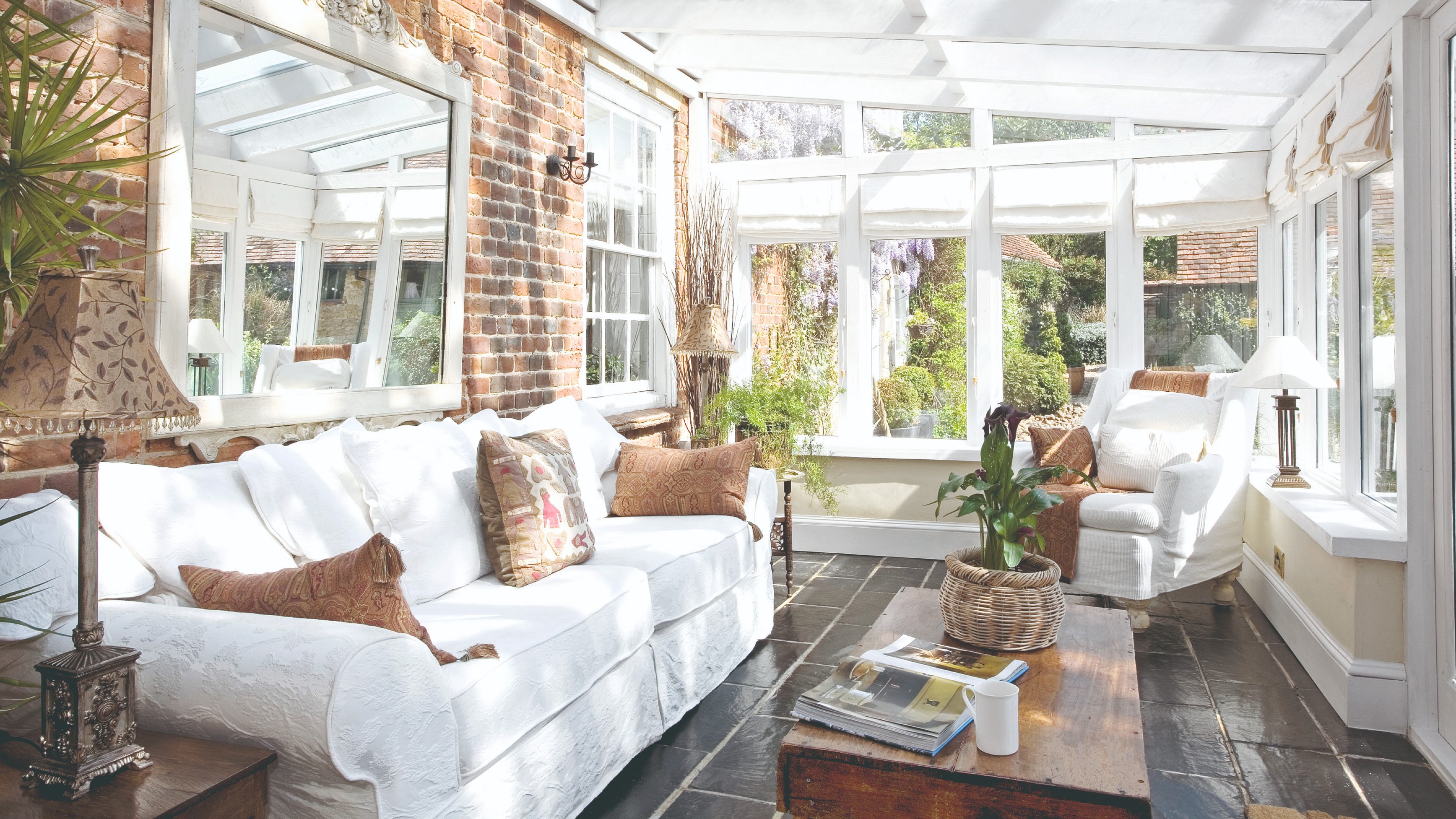 A conservatory with a white sofa and matching armchair and the windows covered with Roman blinds