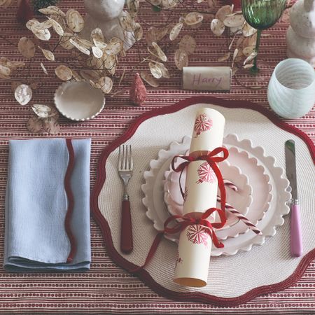 A Christmas table setting with a scalloped placemat and napkin and ribbon-decorated cracker