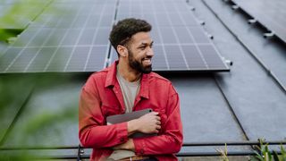 A young worker leaning against a solar panel to represent climate perks, while holding a tablet and wearing a red jacket.