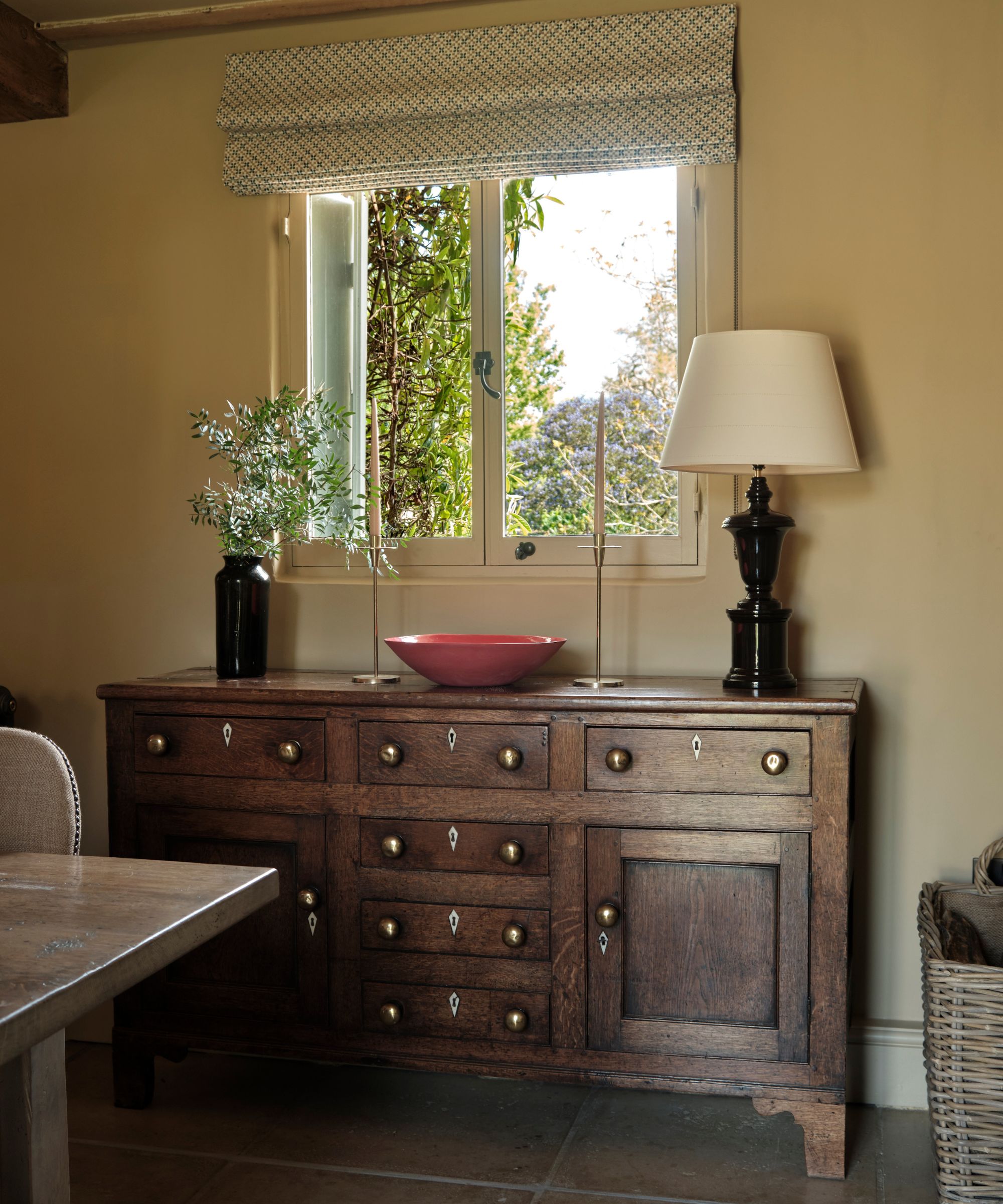 Antique oak sideboard with drawers and cupboards in a traditional kitchen, styled beneath a window with a table lamp, candlesticks and decorative accessories.