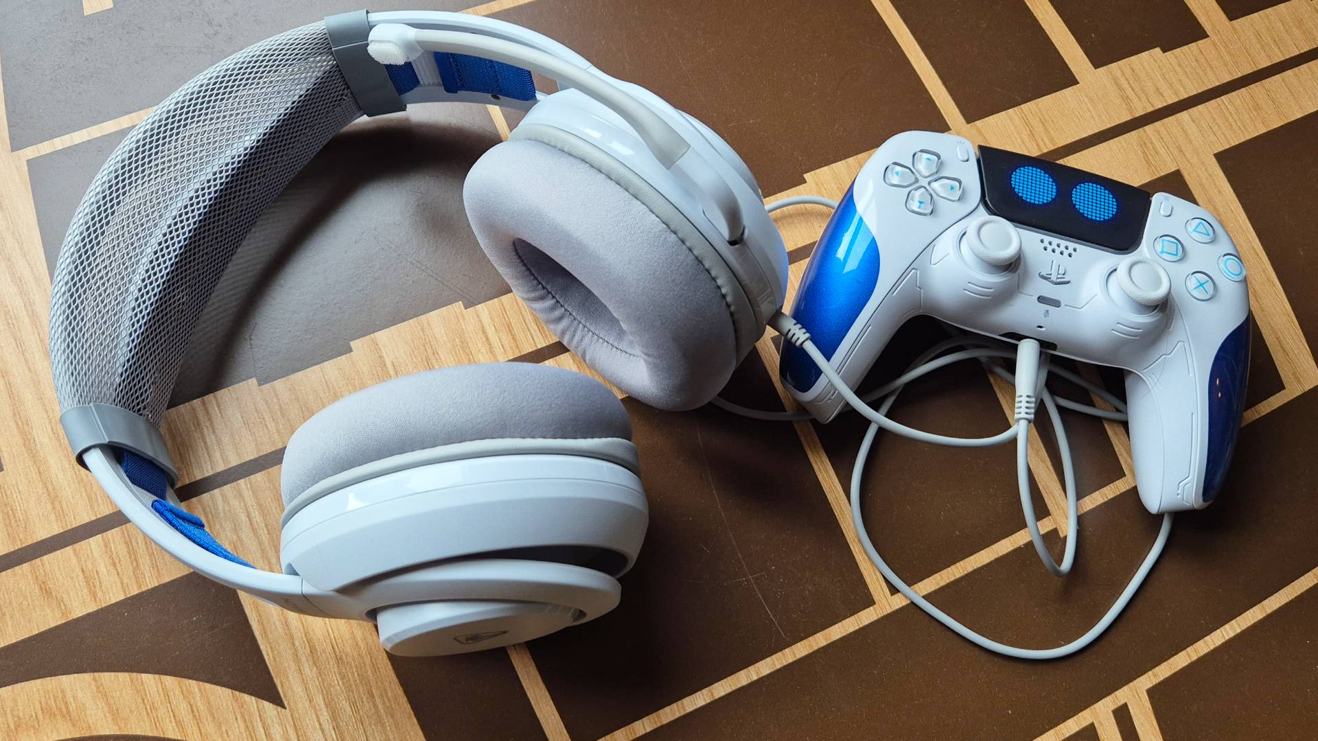 Photo of a white headset sitting on a wooden desk next to a PS5 DualSense controller.