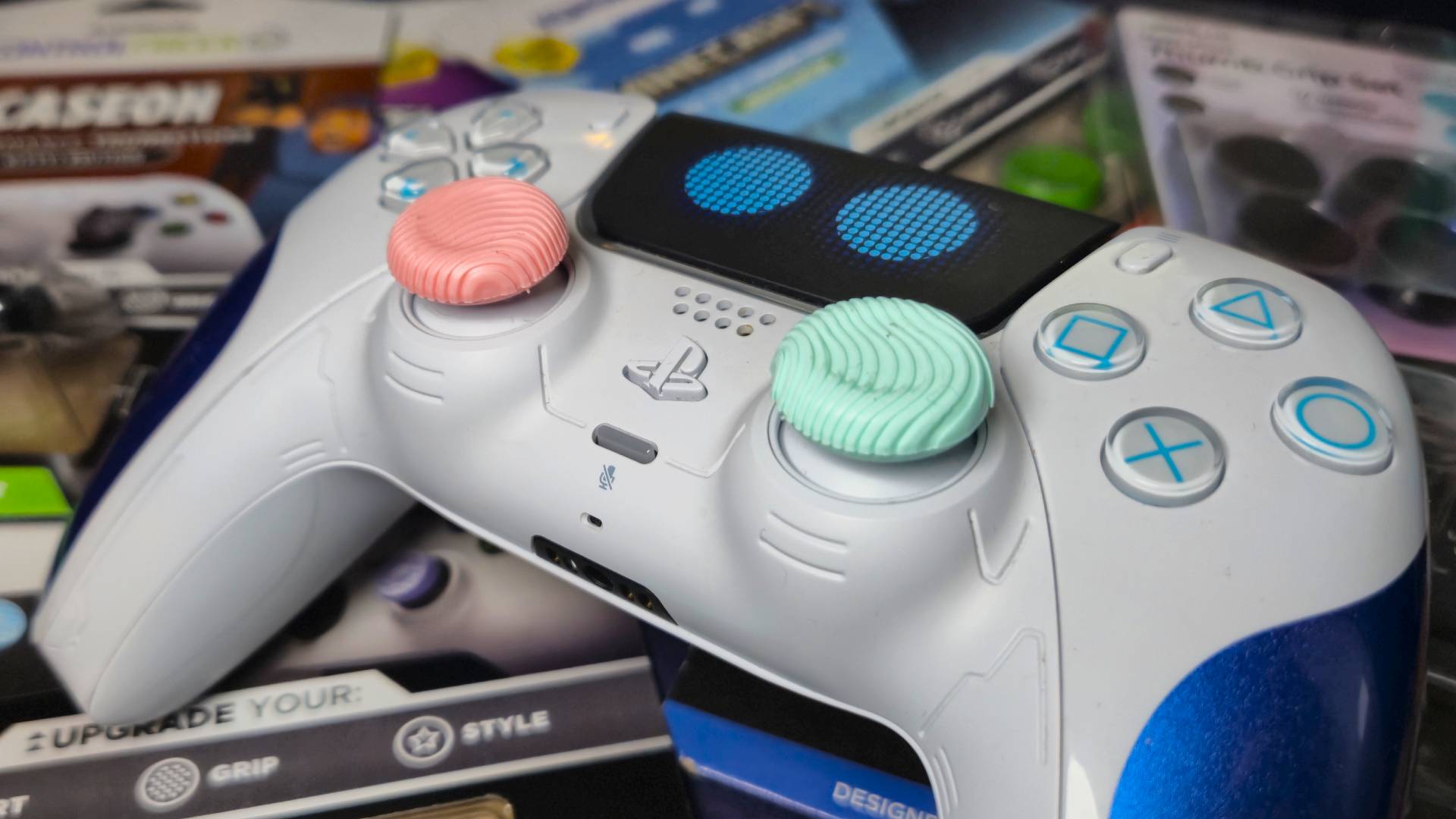 Photo of the Astro Bot PS5 DualSense controller with a pair of pink and green thumb grips. The controller is sitting on a pile of thumb grips packaging.