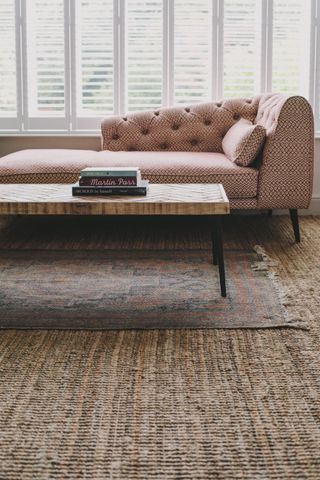 A naturally lit hotel room features a wooden coffee table, rattan carpet, a colorful rug, and a sculptural chaise lounge sofa.