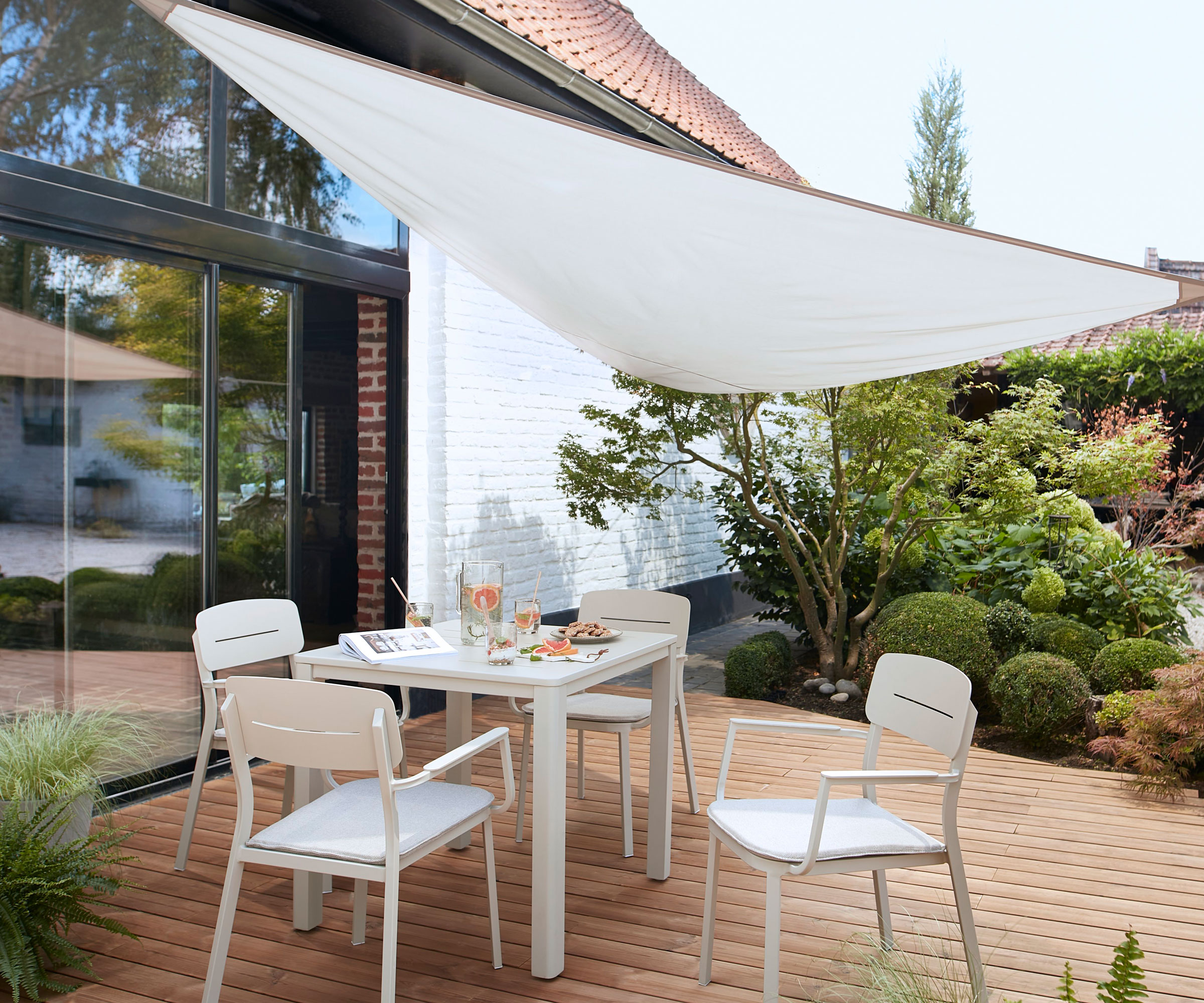 shade sail covering deck with garden furniture set, planting and small tree, and patio doors in background