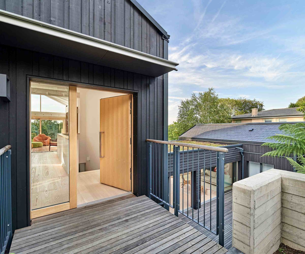 The entrance to a black timber-clad new build, built by Baufritz, featuring a large timber front door and a small bridge over a courtyard above