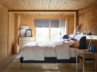 A wooden walled bedroom with bed in center of room, blue bed, white bedding, wooden side table, blinds and drapes