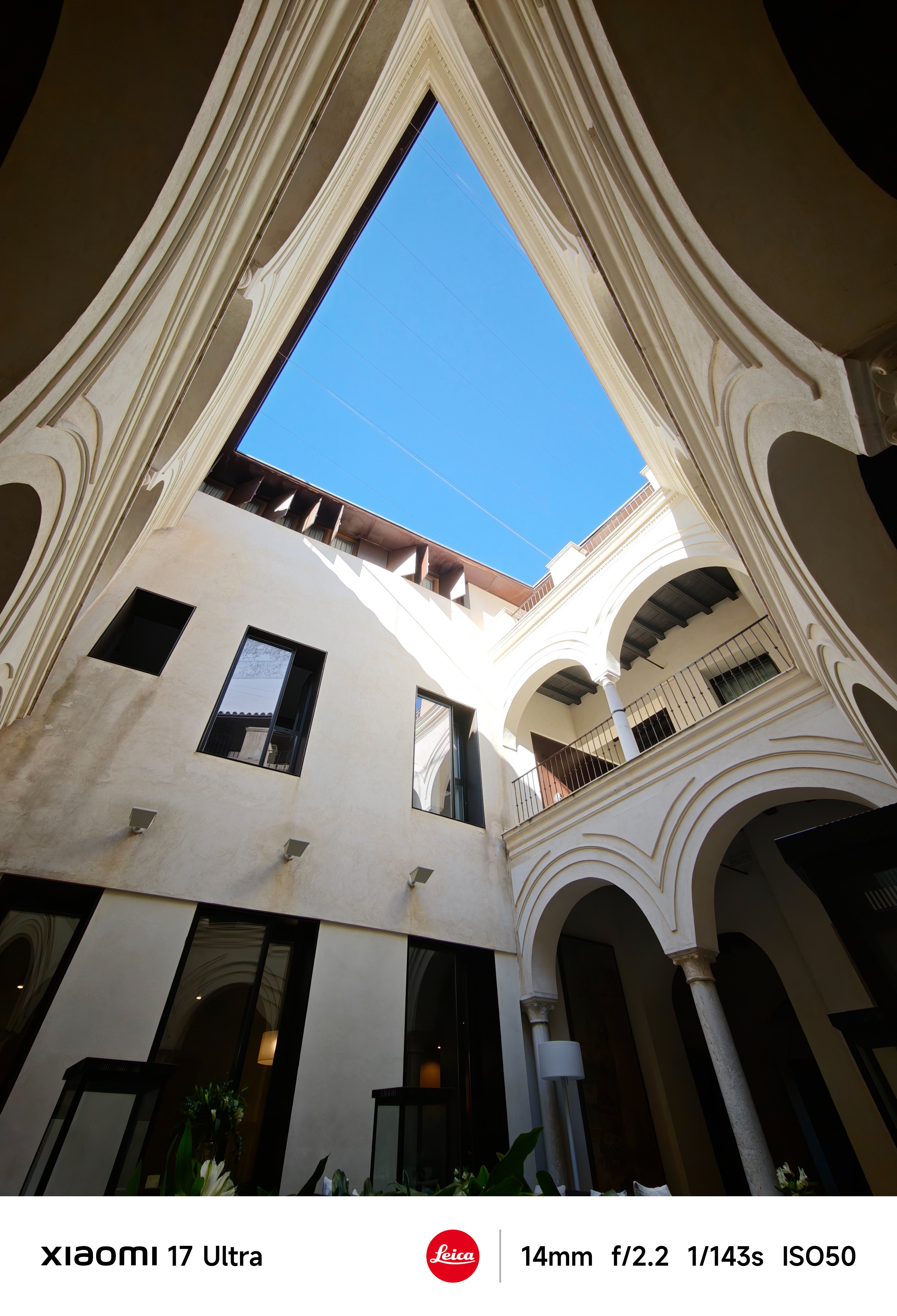 Upward view from a shaded courtyard framed by ornate cream arches, revealing a bright blue sky above.