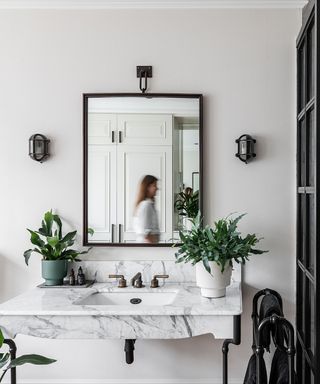 A white bathroom with a black square mirror and black wall sconces either side, a marble sink with two leafy plants on it, and a black towel rack