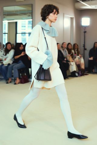 Altuzarra model wearing a cream sweaterdress with white tights and black shoes at the F/W 24 show.