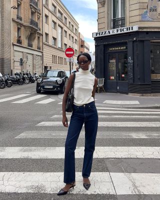 Influencer stands in Paris wearing white shell top and dark wash jeans, taking outfit photo.