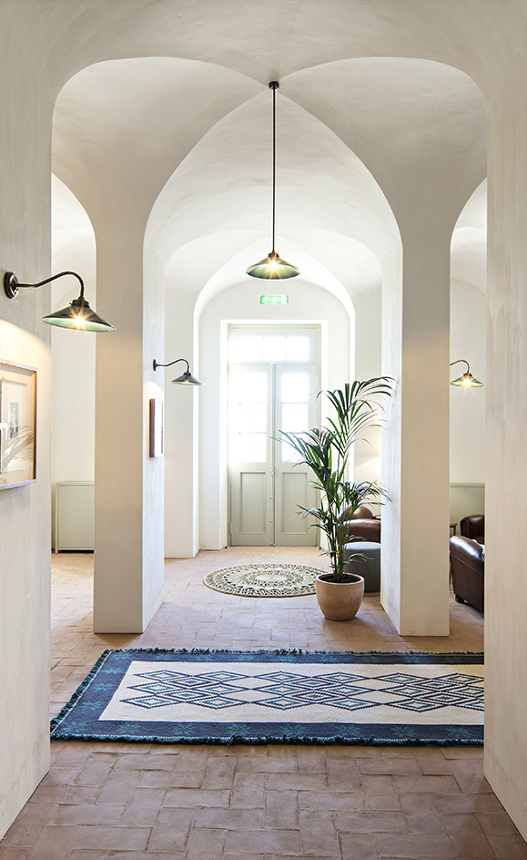 A hallway in the São Lourenço do Barrocal hotel. White walls with a domed ceiling, arched passages to the left and right, and a door to the farthest wall in the center. The floor is covered with rugs in different shapes, in gray, blue, and white colour.