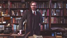 Mark Gattis surrounded by books, leans on a desk with a typewriter next to him