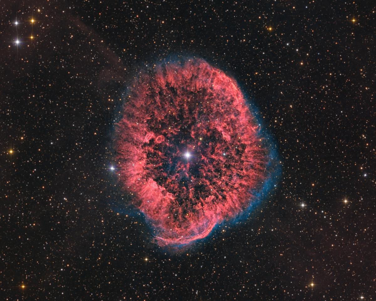 A bright white star sits in the center of a nebula of multi-layered red gasses. Bright spots of distant stars and galaxies surround the red, circular burst.