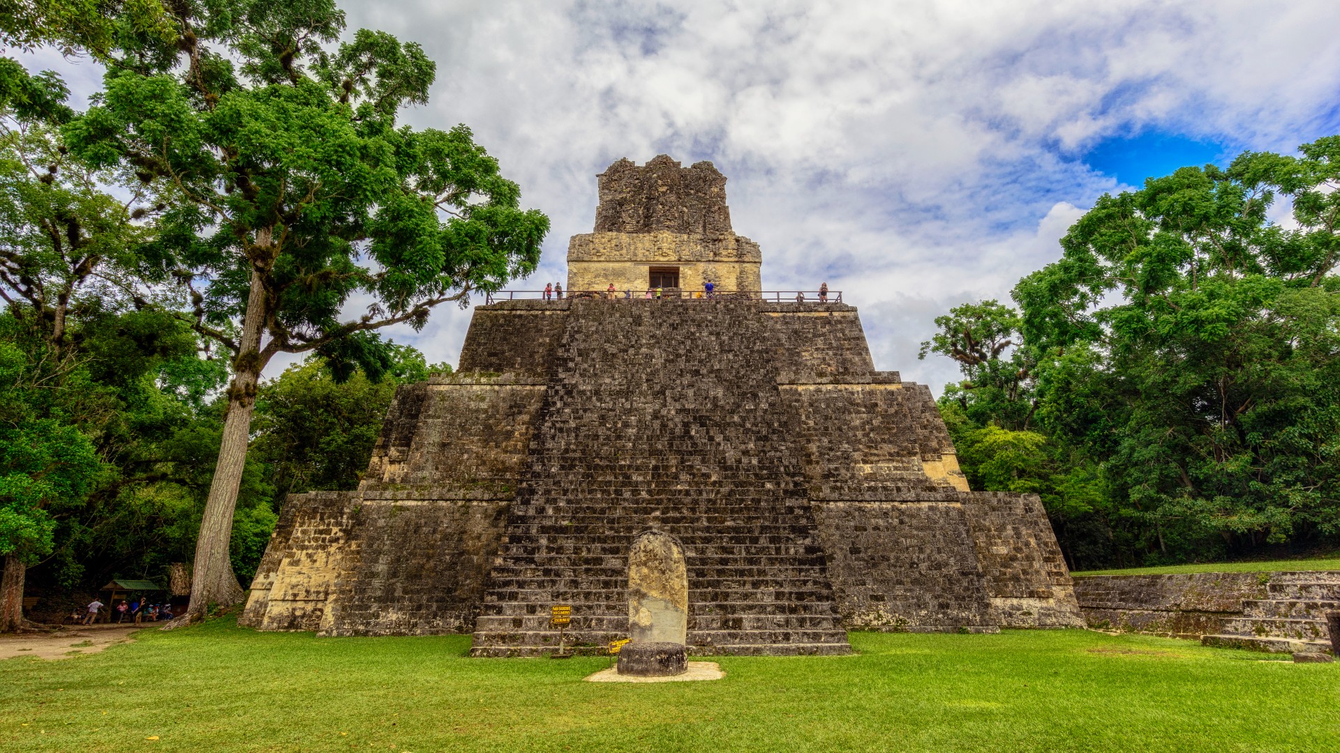 The Masks Temple in the Great Square inside the archeological site of Tikal, Guatemala. It is a short step pyramid (only 4 levels tall). It has a wide staircase in the center leading to an open doorway at the top. At the bottom in front of the stairs there is a large tombstone-shaped stone. There is a large tree to the left which is just as tall as the step pyramid.