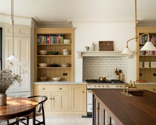 kitchen diner with yellow cabinetry and counter cabinets, metro tile backsplash behind stove, wooden kitchen island with sink, brass faucet, round mahogany table with black chairs, black painted window frames, enamel and brass pendant above island, wood floorboards